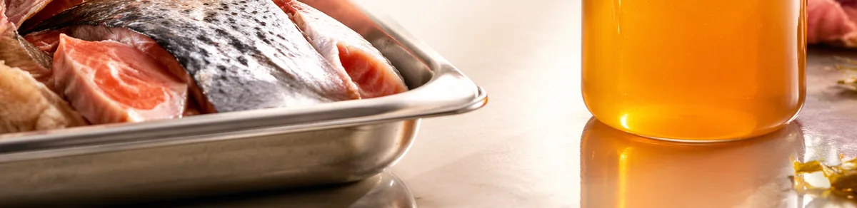 Fish frames and trimmings in a stainless tray beside a jar of clear amber protein hydrolysate